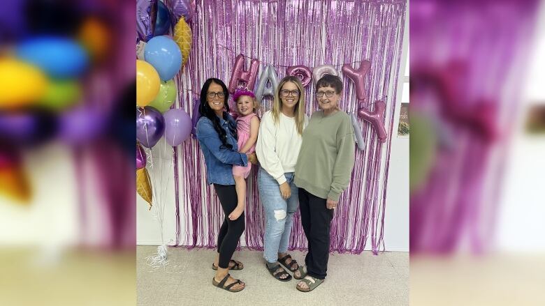 Three women can be seen standing in front of birthday party decorations. One of them is holding a toddler in her arms. 