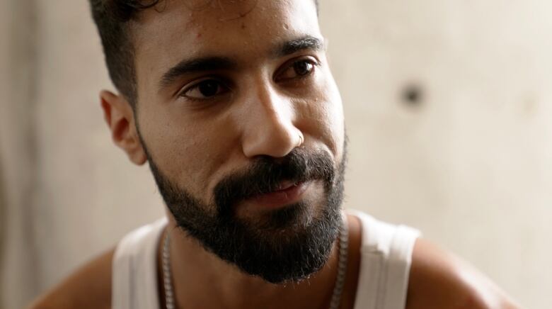 A close-up image of a man with facial hair wearing a white tank top.