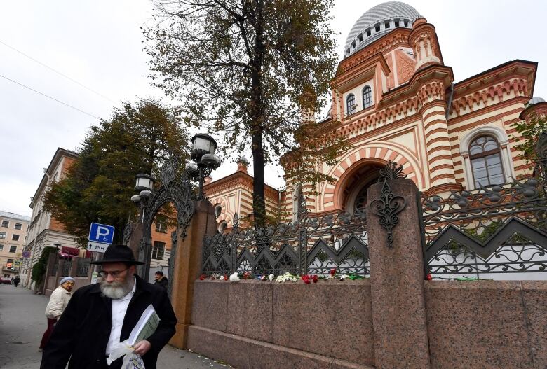 A bearded man in a dark hat is shown on a sidewalk in front of an ornate building that appears to be a house of worship. Flowers are shown on the ledge of a fence outside the building.