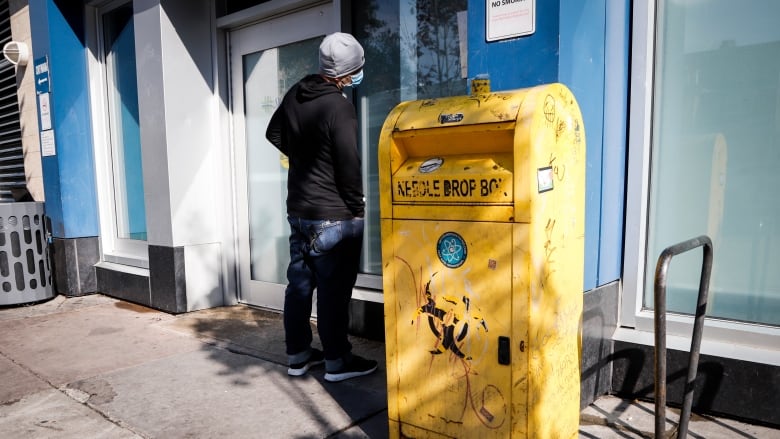 A man wearing a toque and a mask stands outside a building where there is a bin outside that reads 