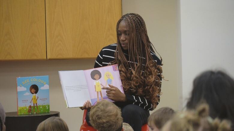 A woman sitting in front of a group of children with an open book in her arms, displaying the inside pages.