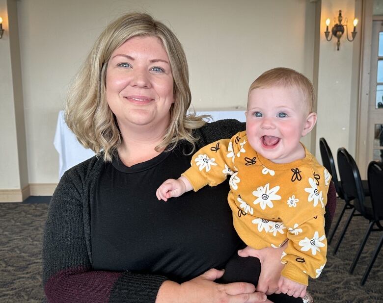 A blonde woman in a black sweater holds a smiling baby.