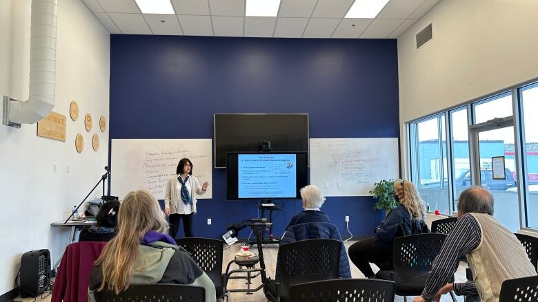 A woman speaks to a gathered audience next to a whiteboard and power point presentation. 