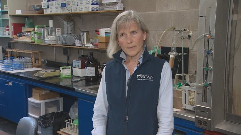 A woman wearing a blue vest stands in a room, with lab equipment in the background.