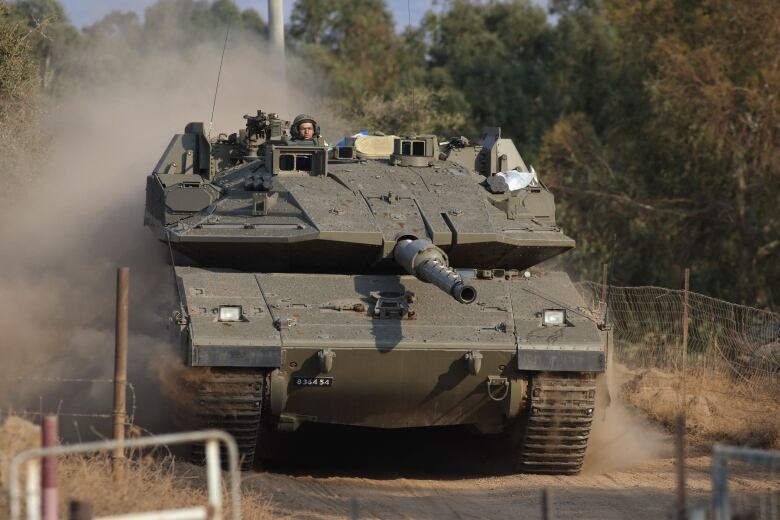 A helmeted person is shown atop a military tank travelling on a dirt path.