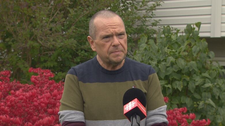 A bald man wearing a green and grey sweater stands in a garden. He's speaking in front of a microphone, and is directly in front of a large rose bush.