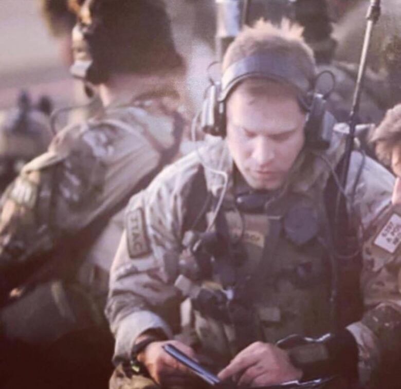 A man in camouflage with a headset is shown looking at a notebook.