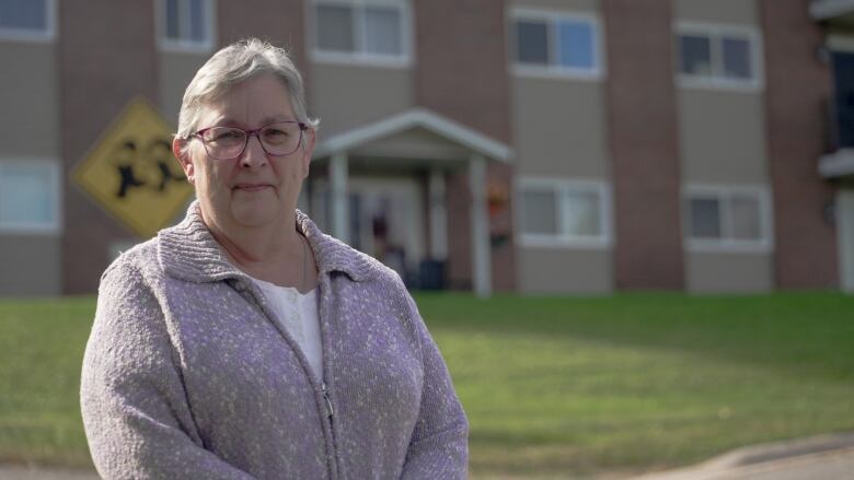 An older lady with short grey hair, wearing a lilac zip-up sweater, standing in front of an apartment building.