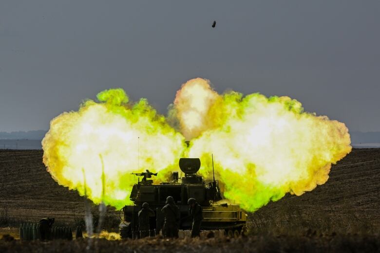 Three figures stand near a piece of military machinery as a huge explosive cloud, tinted green, spreads outward. A shell can be seen in the sky.