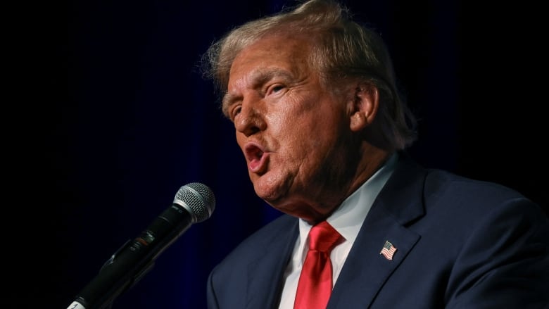 yellow haired man with fake tan and wearing a red tie speaks at podium