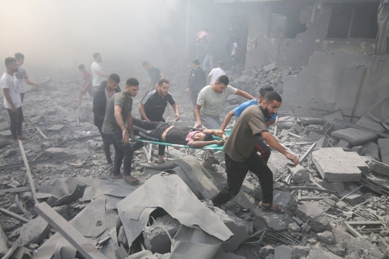 People carrying an injured person in a stretcher, amid rubble and smoke. 