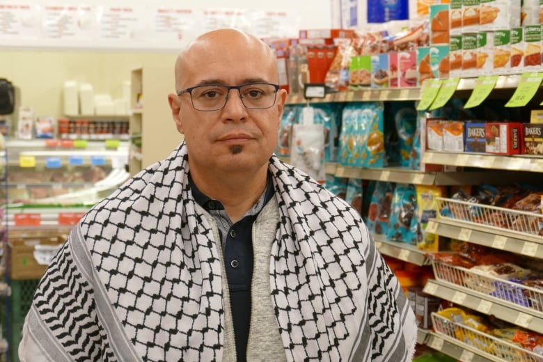 A man, wearing glasses and a black and white shawl draped over his shoulders, stands in front of a grocery store shelf lined with food.