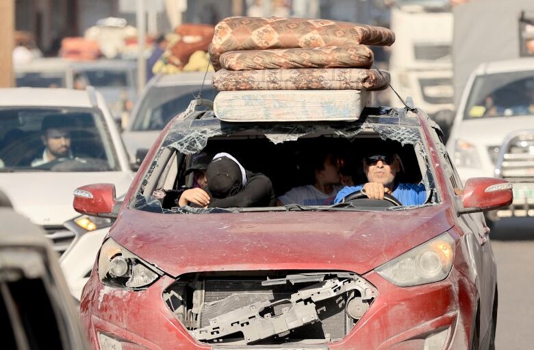 People in a burgundy sedan with the windshield and grill blasted out and a stack of mattresses on top