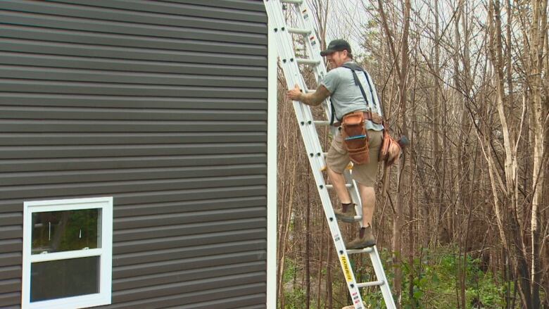 A man is on a ladder. It's on the side of a building with dark grey siding. Behind him are burnt trees. 