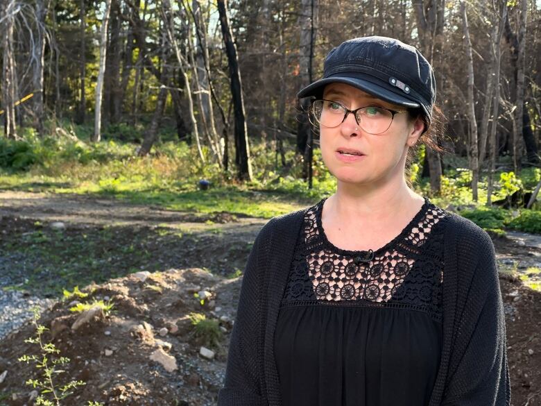 A woman wears a black outfit and black ball cap. She also wears glasses. Behind her is a vacant lot with burnt trees.