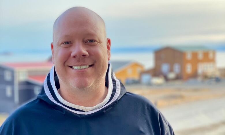 A bald man in a blue pullover smiles at the camera in a photo taken outside. 