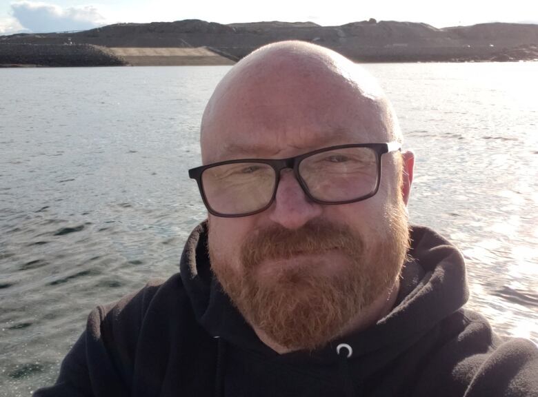 A bald man with a beard and glasses takes a photo outside. 