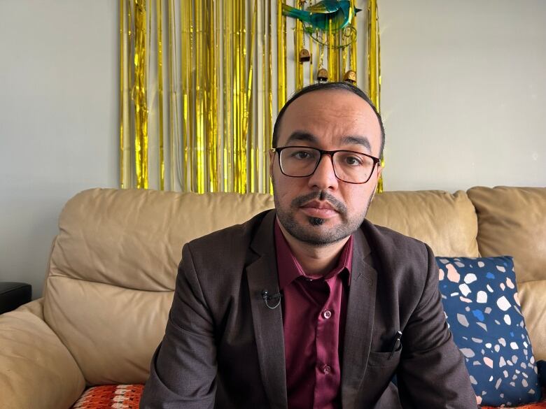 A bald man in a suit sits against a wall with goldens streamers.