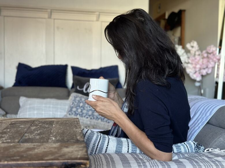 A woman sits on a couch holding a mug, seen in profile with her dark hair covering her face