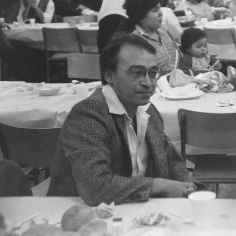 In a black and white photo, a man is shown sitting at a long dining table in the outdoors, with other people in the background.