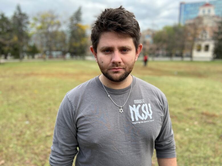 A man in a grey shirt wearing a Star of David necklace stands on a grassy lawn.