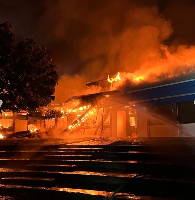 A stream of water from a hose aimed at a large fire burning a building at night.