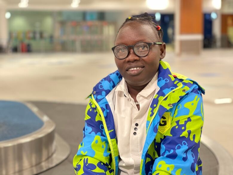 A woman in a bright green and blue jacket poses on a luggage carousel at Halifax Stanfield International Airport. 