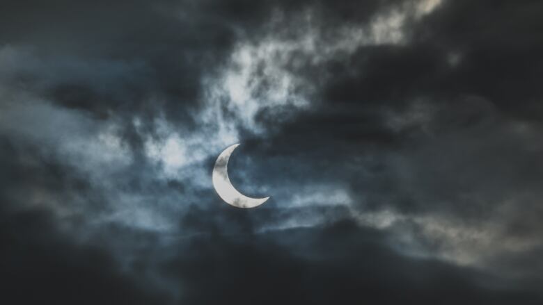 A solar eclipse is shown, with the moon nearly fully blocking out the sun on a cloudy day.