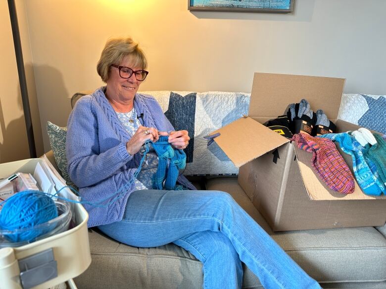 A woman is shown sitting on a couch knitting a pair of blue socks beside a blue ball of yarn and other pairs of socks she is donating to shelters.