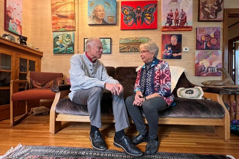 A man with grey hair wearing a grey sweater sits on a wooden couch next to a woman with short grey hair, wearing glasses and a print blouse, surrounded by paintings on the wall.