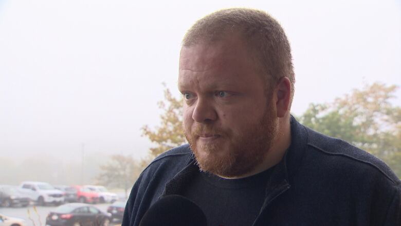 A man with a buzz cut and red beard stands in front of a parking lot.