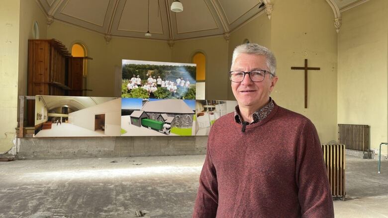 Man smiling inside a run down church, with a decrepit concrete floor and yellow walls. A wooden cross hangs one of the walls behind him.