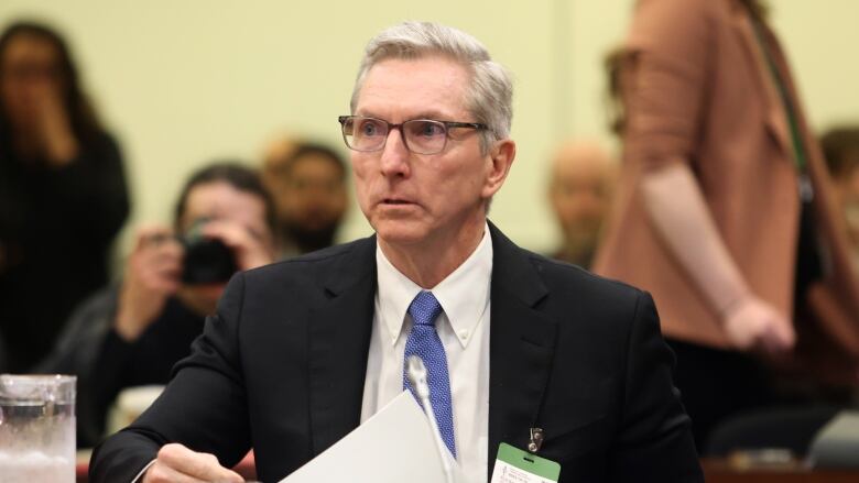 A man with grey hair and glasses is wearing a suit, with a blue tie.