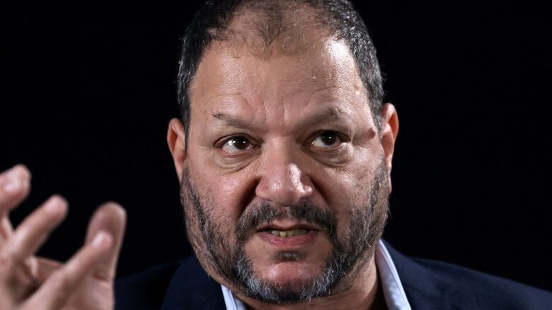 Close up of a balding man with a short, greying beard speaking and gesturing.