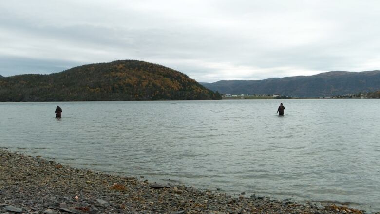 A view of the water's edge with mountains and houses in the background. Two women are walking in the water. 