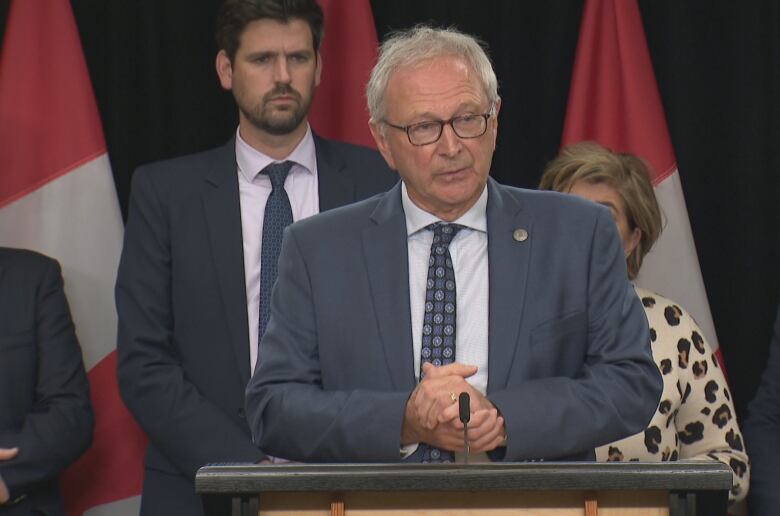 A man in a suit and tie and glasses stands in front of Canadian flags.