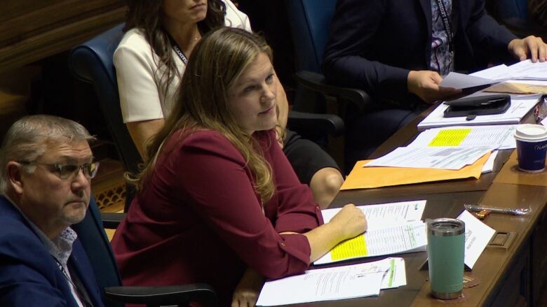 A woman wearing red is seated with her right arm resting on the table in front of her. 