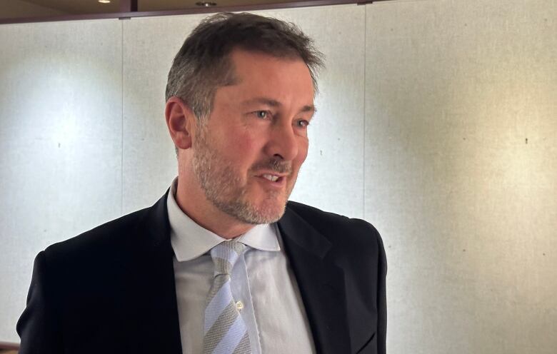 A man with short, greying hair in a black suit with a blank wall in the background.