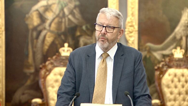 A man with white hair wearing a grey suit with a gold tie stands behind a lectern wearing glasses.