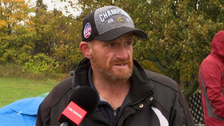 A man wearing a jacket and baseball cap stands on a lawn.