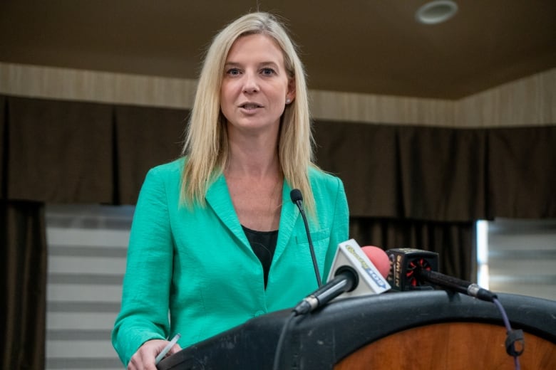 A woman with blonde hair who is wearing a teal-coloured blazer stands behind a podium.