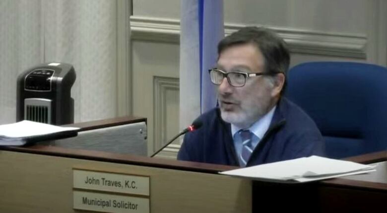 A white man with dark hair, a beard and glasses sits at a desk with a microphone