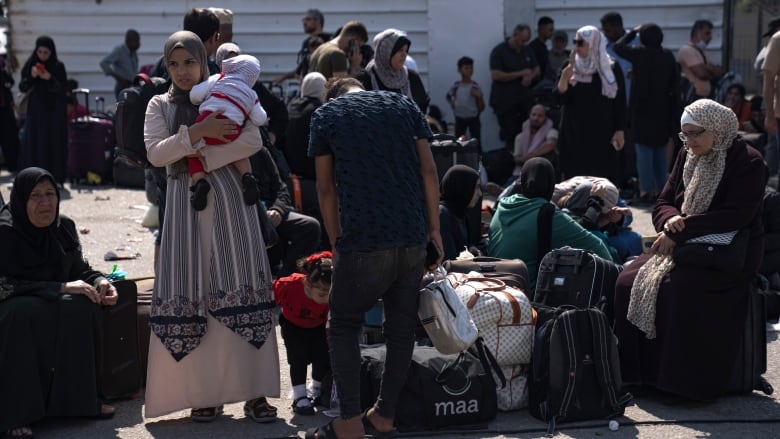 Crowds of men, women and children standing outside with suitcases and bags sitting on the ground. 