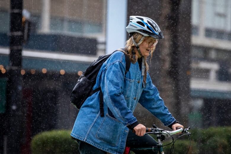 A woman wearing a blue jean jacket and blue bicycle helmet winces during heavy rain.