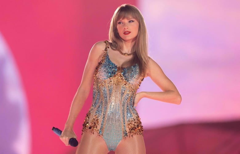 Swift stands on stage, mic in one hand and her other hand on her hip, wearing a body suit with gold and silver rhinestones.