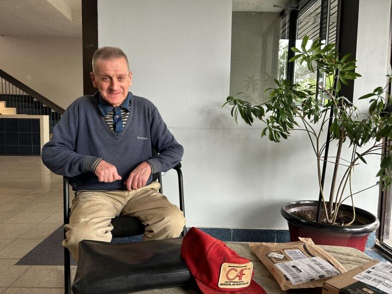 older man in blue sweater, tie, beige pants smiling at camera in lobby of building