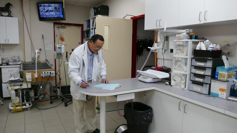 A veterinarian in a white coat is standing in a room with medical equipment surrounding him, while holding a pen to write on papers.