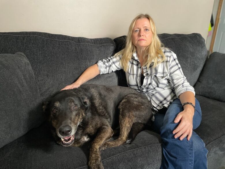 A woman wearing a white shirt with stripes is sitting with her dark-coloured dog on a black couch in her home.