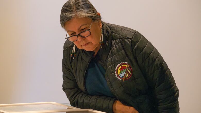 A woman wearing black-rimmed glasses is pictured leaning to look at a document in a glass encasement.