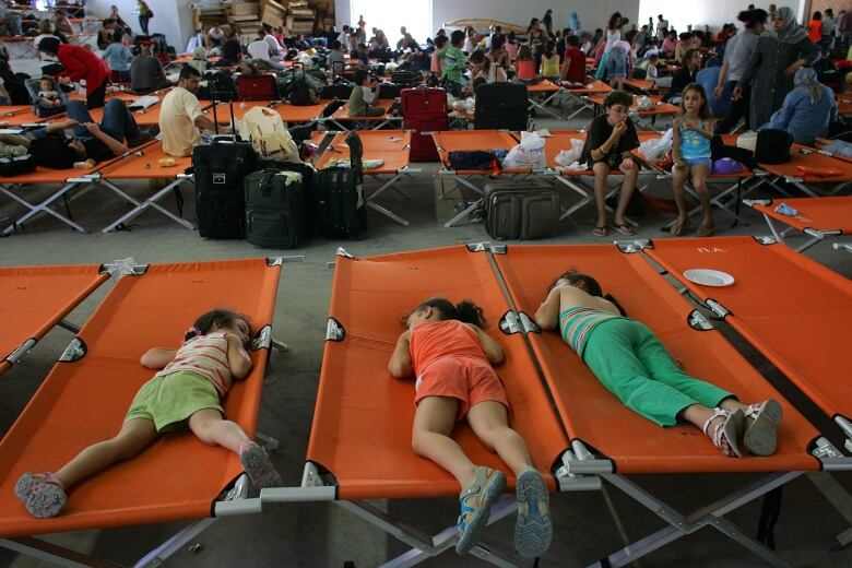 Three children laying on a row of orange cots in front of a room of other people sitting on similar cots. 